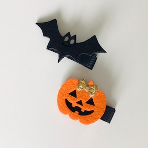 May include: Two Halloween-themed hair clips. One is a black felt bat with a small white face. The other is an orange felt pumpkin with a black face and a gold glitter bow.