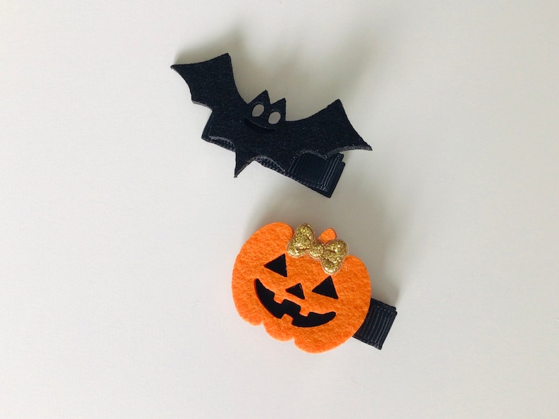 May include: Two Halloween-themed hair clips. One is a black felt bat with a small white face. The other is an orange felt pumpkin with a black face and a gold glitter bow.