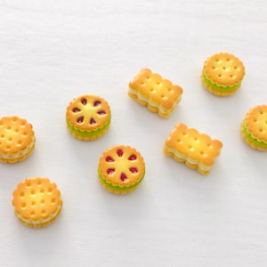 May include: A selection of miniature decorative biscuits. The biscuits are golden brown with a cream filling, and some have a red fruit filling. The biscuits are round and rectangular, arranged on a white surface.