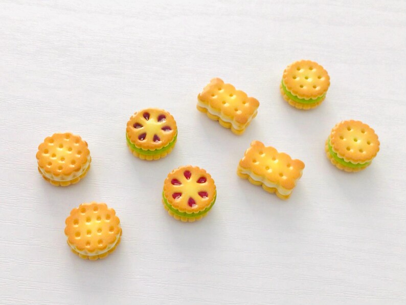 May include: A selection of miniature decorative biscuits. The biscuits are golden brown with a cream filling, and some have a red fruit filling. The biscuits are round and rectangular, arranged on a white surface.