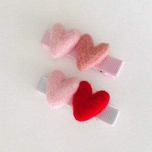 May include: Two hair clips with felt hearts. One clip has three hearts: pink, light pink, and red. The other clip has two hearts: pink and light pink.