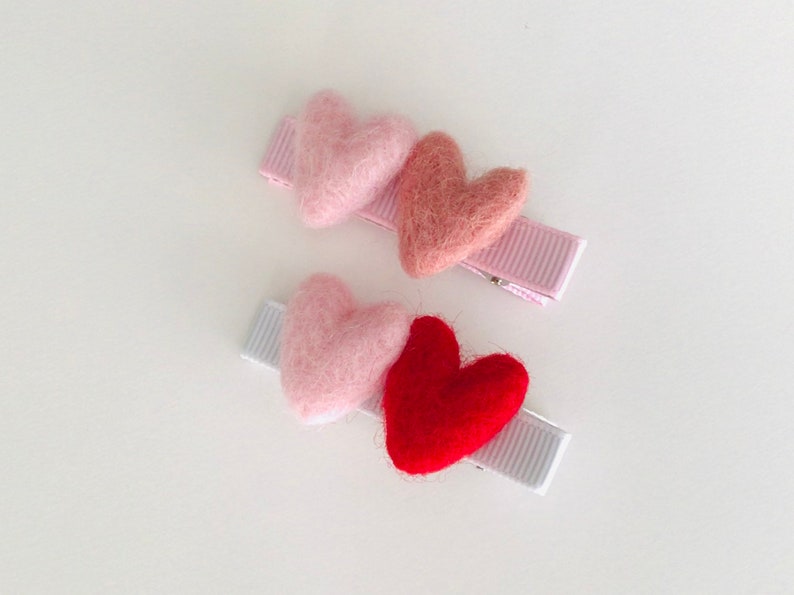May include: Two hair clips with felt hearts. One clip has three hearts: pink, light pink, and red. The other clip has two hearts: pink and light pink.