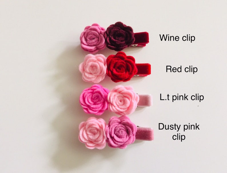May include: Four pairs of felt flower hair clips in various shades of pink and red. The clips are arranged vertically from darkest to lightest, with the top clip labeled "Wine clip", the second clip labeled "Red clip", the third clip labeled "L.t. pink clip", and the bottom clip labeled "Dusty pink clip".