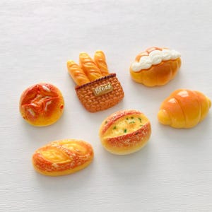 May include: An assortment of miniature bread items, including baguettes in a basket, a croissant, and various rolls. The bread has a golden-brown color with detailed textures, set against a white background.