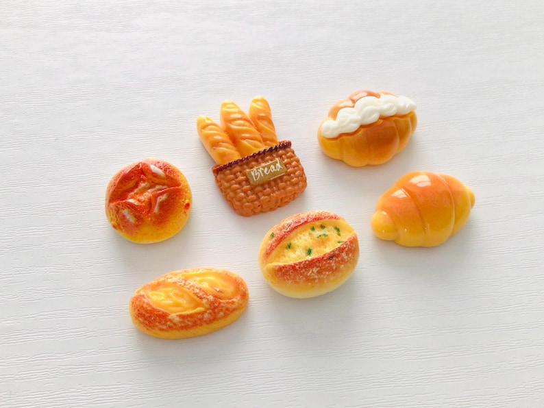 May include: An assortment of miniature bread items, including baguettes in a basket, a croissant, and various rolls. The bread has a golden-brown color with detailed textures, set against a white background.