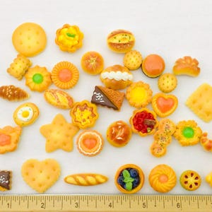 May include: A selection of miniature baked goods, including cookies, pastries, and bread, in various shapes and sizes. The assortment features a star-shaped cookie, a heart-shaped cookie, and a croissant. A ruler is at the bottom, measuring in inches (approx. 2.54 cm).