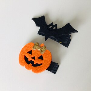 May include: Two Halloween-themed felt hair clips. One is a black bat with a gold glitter bow. The other is an orange pumpkin with a gold glitter bow and a jack-o'-lantern face.