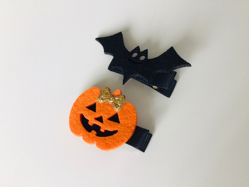 May include: Two Halloween-themed felt hair clips. One is a black bat with a gold glitter bow. The other is an orange pumpkin with a gold glitter bow and a jack-o'-lantern face.