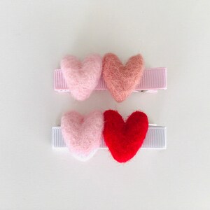 May include: Three felt heart hair clips. The first clip has a pink heart, the second clip has a light pink heart, and the third clip has a red heart.