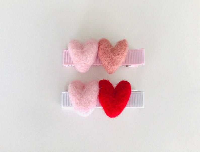 May include: Three felt heart hair clips. The first clip has a pink heart, the second clip has a light pink heart, and the third clip has a red heart.