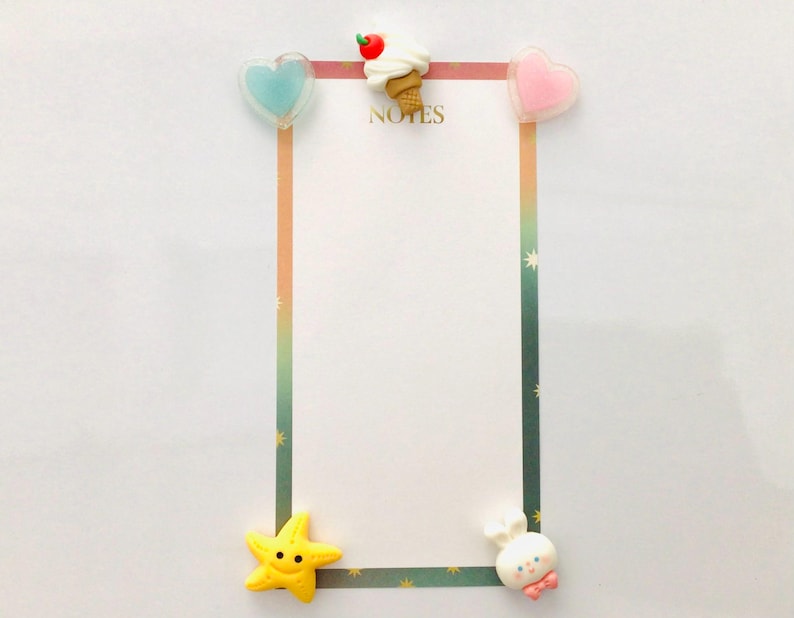 Peut inclure: Un bloc-notes blanc avec une bordure color&eacute;e et le mot "NOTES" en haut. Orn&eacute; d'&eacute;l&eacute;ments d&eacute;coratifs : un c&oelig;ur bleu, une glace, un c&oelig;ur rose, une &eacute;toile de mer jaune et un personnage blanc avec un n&oelig;ud rose.