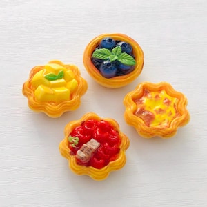 May include: Four miniature tarts with yellow, scalloped edges. One tart is topped with blueberries and a sprig of mint. Another has yellow mango chunks. The third has a red topping with a small piece of brown food. The last tart has a yellow and orange topping.