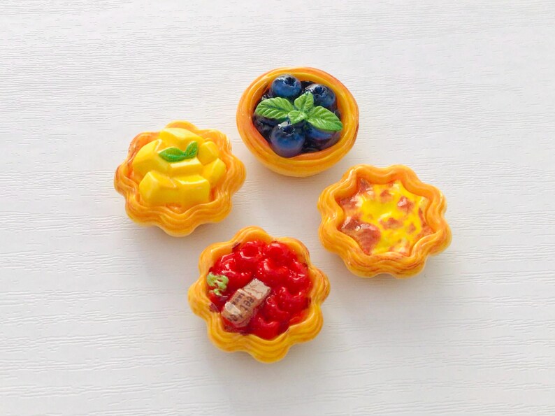 May include: Four miniature tarts with yellow, scalloped edges. One tart is topped with blueberries and a sprig of mint. Another has yellow mango chunks. The third has a red topping with a small piece of brown food. The last tart has a yellow and orange topping.