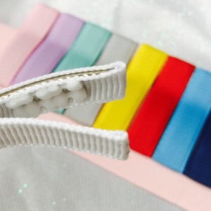 May include: A white hair clip with a row of white pom-poms on the top. The clip is open and is positioned above a rainbow of colorful ribbon.