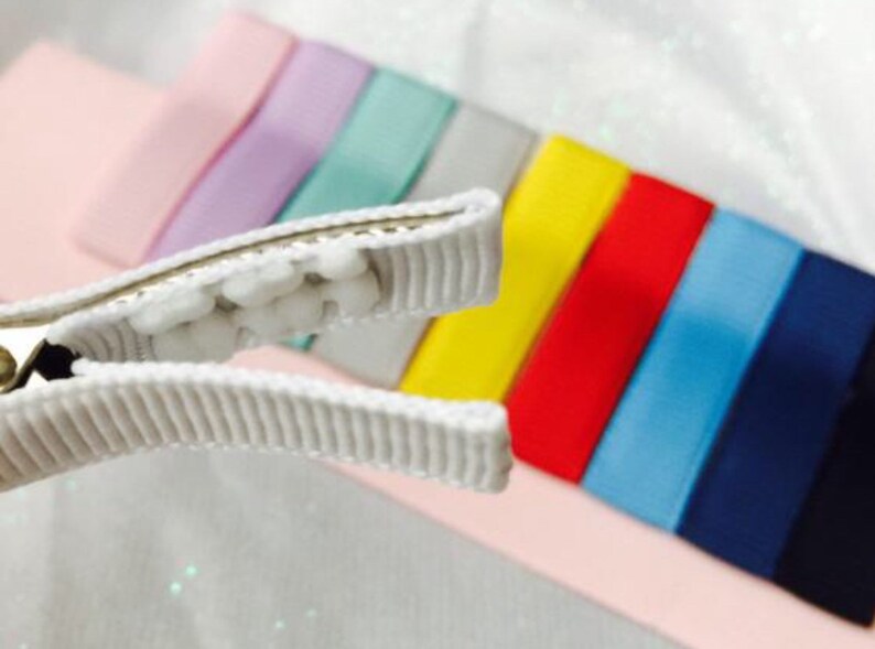 May include: A white hair clip with a row of white pom-poms on the top. The clip is open and is positioned above a rainbow of colorful ribbon.