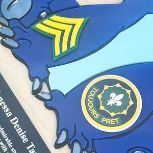 May include: A blue, die-cut emblem with a light blue stripe and a yellow and green chevron. A circular emblem with the words "TOUJOURS PRET" surrounds a green fleur-de-lis. The name "Vanessa Denise Tate" is printed on a black rectangle.