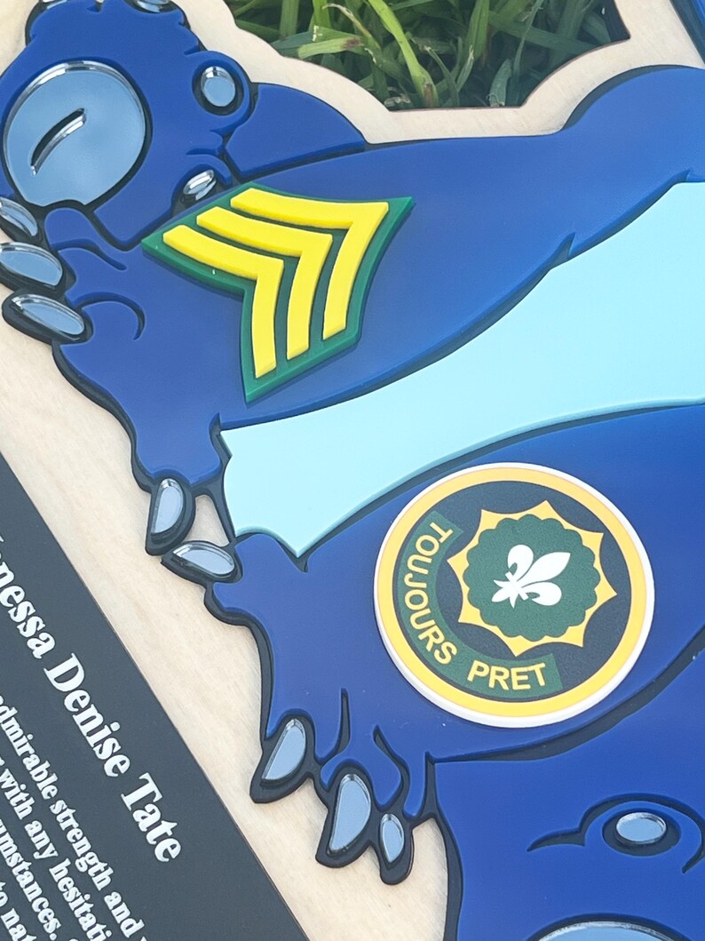 May include: A blue, die-cut emblem with a light blue stripe and a yellow and green chevron. A circular emblem with the words "TOUJOURS PRET" surrounds a green fleur-de-lis. The name "Vanessa Denise Tate" is printed on a black rectangle.