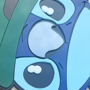 May include: A close-up of a blue, green, and gray cartoon character face. The character has large eyes with white accents and a light blue nose. The design is layered with different shades of blue.