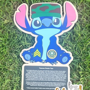 May include: A blue cartoon character with large ears and a camouflage hat. The character is holding a sign with text and a gold glitter signature. The sign reads "Vanessa Denise Tate" and "Ohana means family."