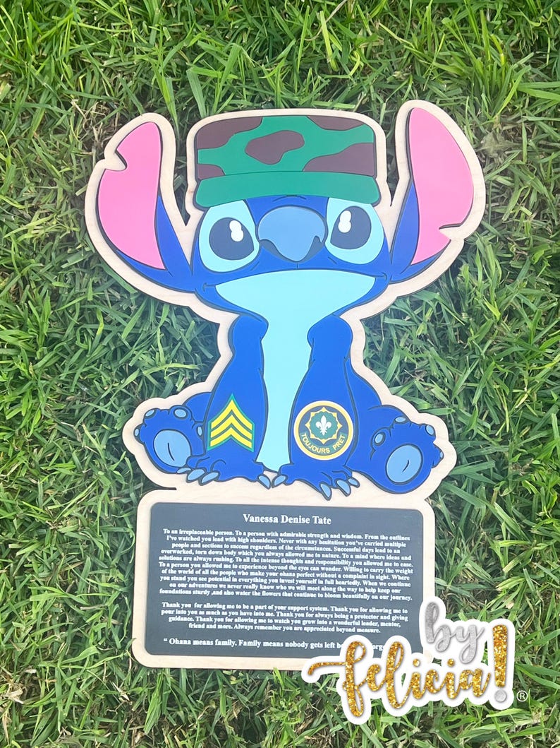 May include: A blue cartoon character with large ears and a camouflage hat. The character is holding a sign with text and a gold glitter signature. The sign reads "Vanessa Denise Tate" and "Ohana means family."