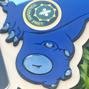 May include: A blue, laser-cut wooden sign with a cartoon-like design. The sign features a circular emblem with the words "TOUJOURS PRET" and a fleur-de-lis symbol. The sign has a light wood base and a blue top layer with reflective accents.