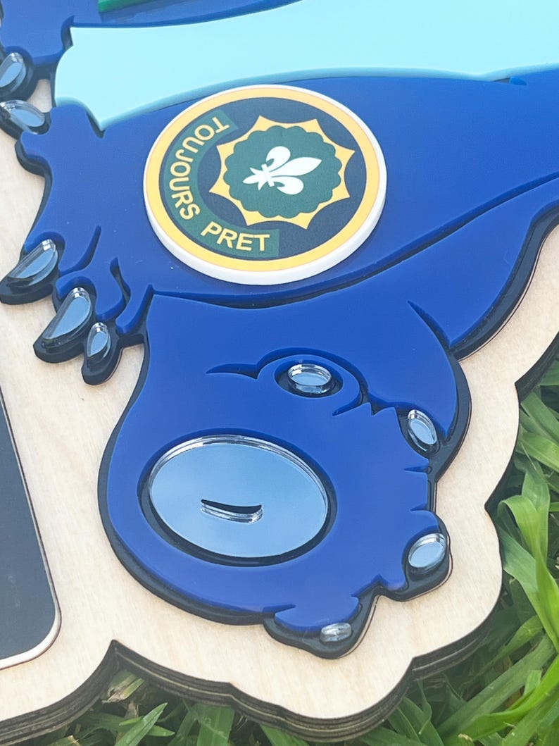 May include: A blue, laser-cut wooden sign with a cartoon-like design. The sign features a circular emblem with the words "TOUJOURS PRET" and a fleur-de-lis symbol. The sign has a light wood base and a blue top layer with reflective accents.