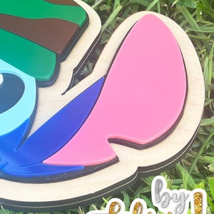 May include: A layered wooden craft featuring a cartoon character's head, with green, brown, blue, and pink accents. The design is set against a background of green grass. The text "by Felicia!" is visible in gold glitter.