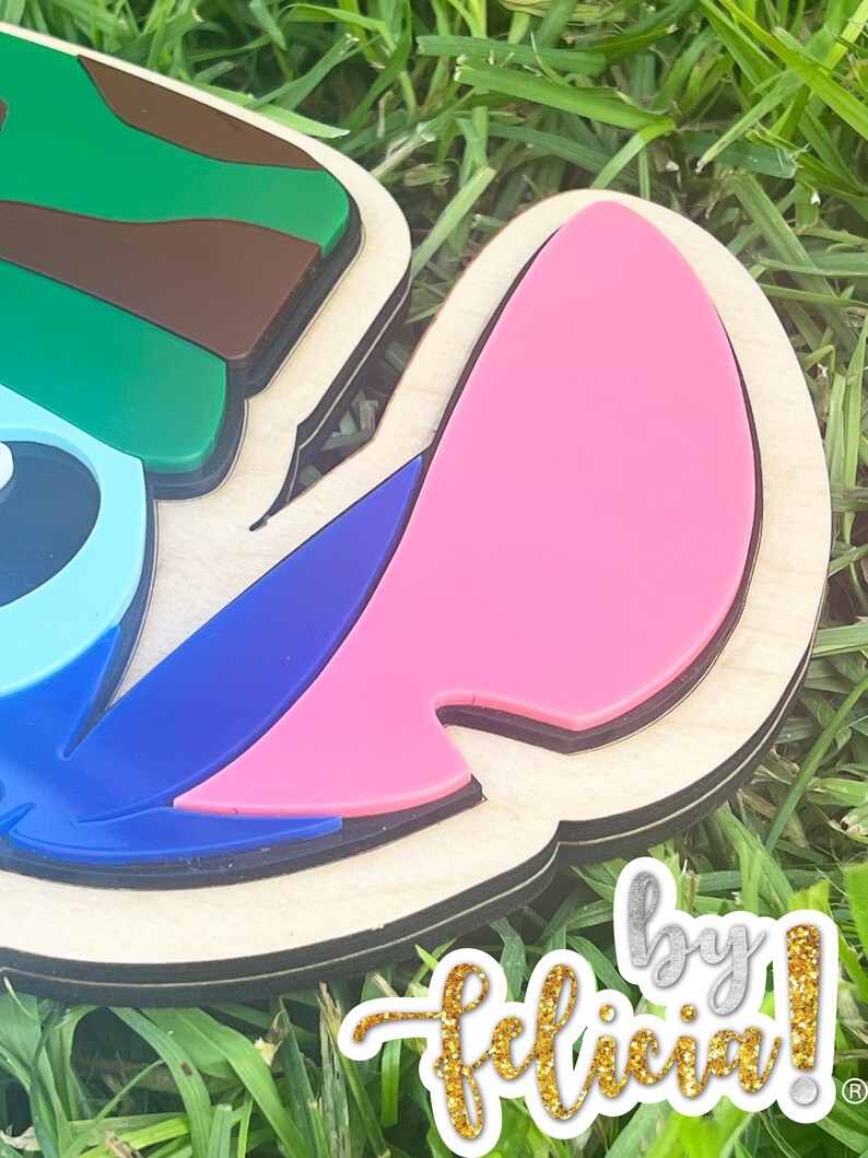 May include: A layered wooden craft featuring a cartoon character's head, with green, brown, blue, and pink accents. The design is set against a background of green grass. The text "by Felicia!" is visible in gold glitter.