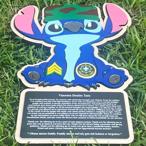 May include: A wooden Stitch character cutout with a camouflage headband, military insignia, and the words "TOUJOURS PRET." A plaque below reads "Vanessa Denise Tate" and includes a quote about family.