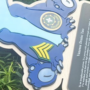 May include: A wooden plaque featuring a blue cartoon-like character with the text "TOUJOURS" and a circular emblem. The plaque also includes text that reads "Vanessa Denise Tate". The character has a light blue background and a yellow and green emblem.