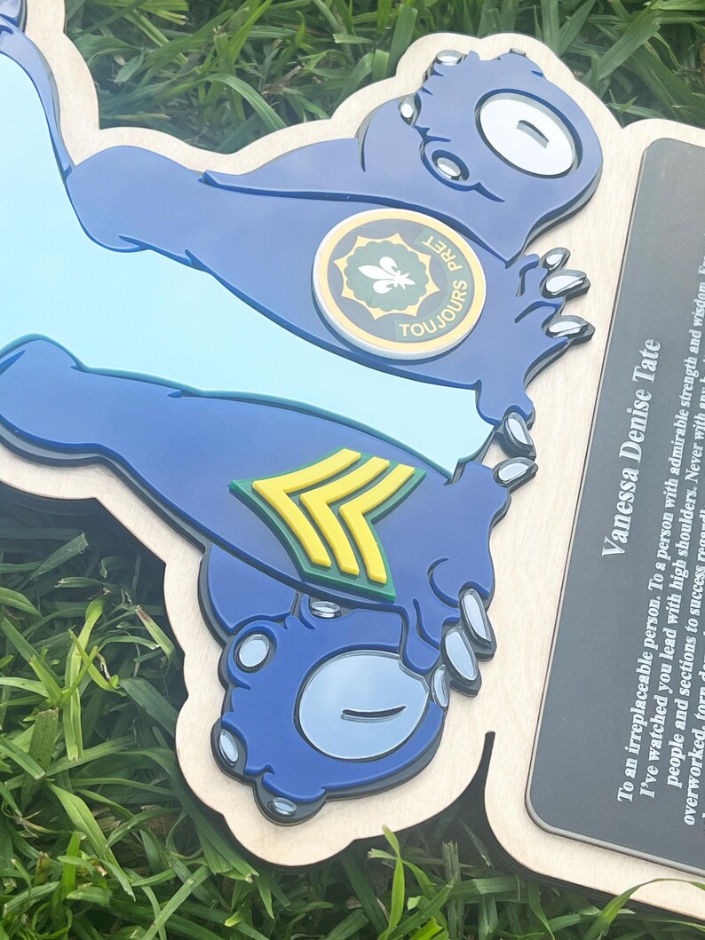 May include: A wooden plaque featuring a blue cartoon-like character with the text "TOUJOURS" and a circular emblem. The plaque also includes text that reads "Vanessa Denise Tate". The character has a light blue background and a yellow and green emblem.