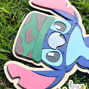 May include: A layered wooden cutout of the character Stitch from Lilo & Stitch. The cutout features blue, pink, green, and brown colors. The text "by Felicia!" is in the bottom right corner.