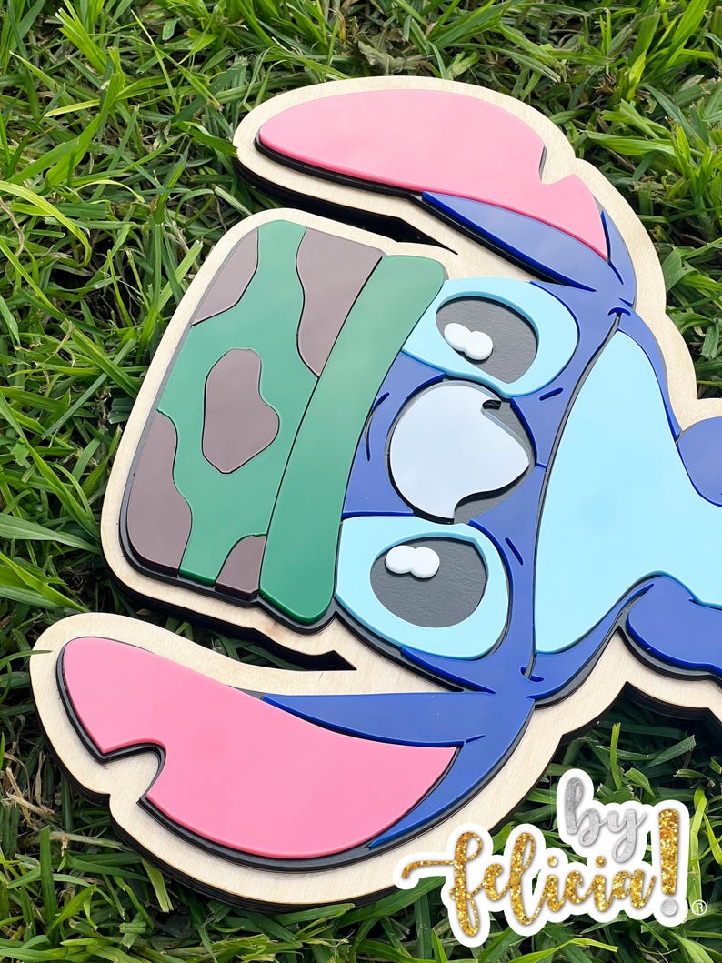 May include: A layered wooden cutout of the character Stitch from Lilo & Stitch. The cutout features blue, pink, green, and brown colors. The text "by Felicia!" is in the bottom right corner.