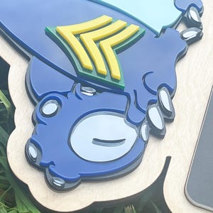 May include: A layered wooden plaque featuring a blue and light blue design with yellow and green accents. The plaque has a unique shape and a glossy finish. The design includes a stylized emblem.