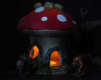 May include: A whimsical mushroom house with a red cap and white spots, featuring two arched doorways with glowing orange light. The house is surrounded by miniature figurines, including a warrior and a creature.