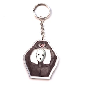 Puede incluir: Un llavero con un anillo y una cadena plateados. El colgante tiene forma hexagonal blanca con un dibujo de una figura haciendo un corazón con las manos. Un pequeño corazón rojo está en el centro.