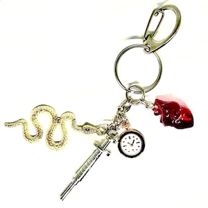 Puede incluir: Un llavero plateado con dijes de serpiente dorada, jeringa, reloj y corazón rojo. La serpiente es detallada. El reloj tiene una esfera blanca y números negros. El corazón es rojo.