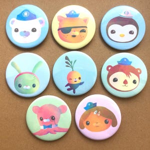 May include: Eight circular pin badges with cartoon animal characters. Each badge has a light blue, yellow, or pink background. The designs include a bear, cat, penguin, bunny, and octopus, all wearing sailor hats. The badges are brightly coloured.