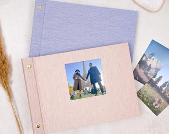 May include: A light purple fabric photo album and a light pink fabric photo album with a square window cutout on the front cover. The pink album features a photograph of two people standing in front of the Eiffel Tower in Paris, France.