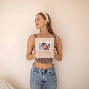 Puede incluir: Un álbum de fotos cuadrado y de color claro con una fotografía de una pareja besándose. El álbum tiene una cubierta texturizada y letras doradas. El álbum lo sostiene una persona que lleva un top marrón y vaqueros azules.