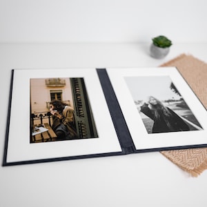 May include: A dark blue, open, bi-fold photo folio featuring two white mat boards. The left side displays a color photograph of a person sitting on a balcony, while the right side contains a black and white portrait photograph of a person.