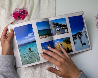 May include: An open photo album displays vacation photos with clear blue water and bright skies. The album is held by a person with manicured nails. Pink flowers and a white textured surface are in the background.