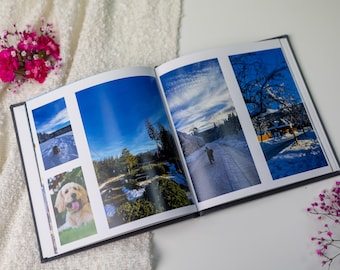 May include: An open photo album with landscape and dog photos. The album is on a white surface with pink flowers. The photos feature blue skies, snowy scenes, and a golden retriever. The album is open, displaying multiple images.