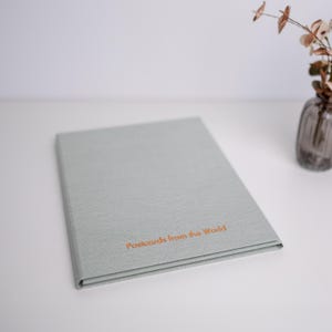 Pode incluir: Um álbum retangular cinza-claro com uma capa de tecido texturizado. O texto "Postcards from the World" está impresso em laranja na parte central inferior da capa frontal. O álbum está posicionado sobre uma superfície plana ao lado de um vaso de vidro.