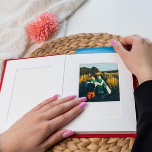 Könnte beinhalten: Ein rotes Fotoalbum mit weißem Passepartout und einem Foto von zwei Personen auf einem Feld. Das Album ist geöffnet, und eine Hand hebt eine blaue Lasche an, um das Foto zu entfernen. Ein rosa Pompon und eine gewebte Matte befinden sich im Hintergrund.