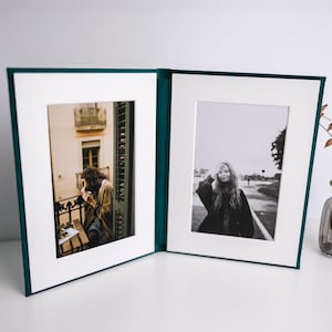 May include: An open, teal-colored photo album with two white-matted picture frames. One frame holds a color photo of a person on a balcony, the other a black and white photo of a person outdoors.