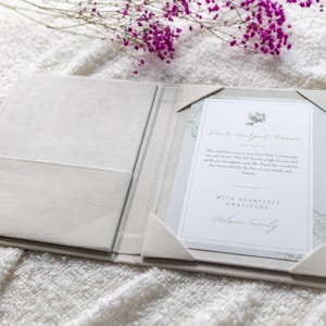 May include: A light beige, fabric-covered presentation folder featuring a pocket on the left and corner tabs on the right holding a white card. The card displays text including "First Holy Communion" and "WITH HEARTFULLY GRATITUDE" in gold and black ink.