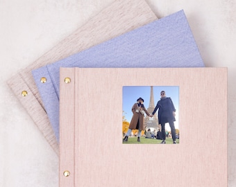 May include: Three rectangular photo albums with fabric covers in beige, light blue, and pink. The top pink album features a square inset photograph of two people holding hands in front of the Eiffel Tower. Each album has two gold metal screw posts.