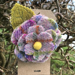 May include: A tweed fabric flower brooch with a yellow central bead and a green leaf. The flower is made of purple, pink, and orange tweed. The word "bloom" is printed on the tag attached to the brooch.