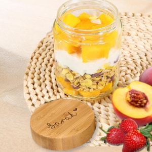 Op de afbeelding: Een glazen pot gevuld met lagen granola, yoghurt en in blokjes gesneden mango. Een houten deksel met de naam "Sarah" gegraveerd ligt naast de pot. Verse aardbeien en een gesneden perzik zijn ook zichtbaar.